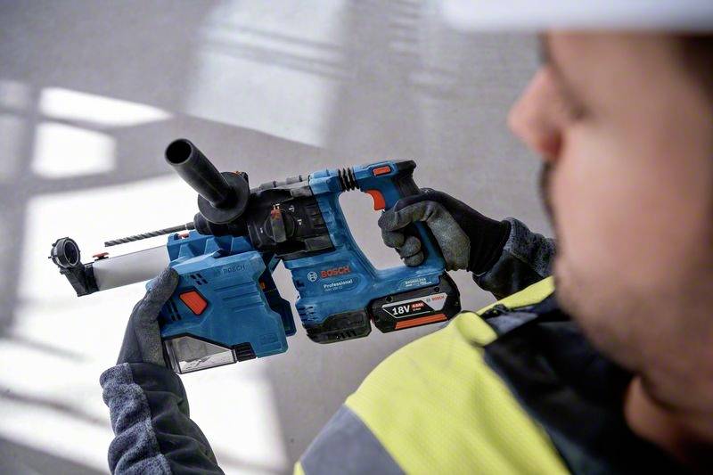 Dělník drží modrý příklepový vrtací kladivo s odsáváním prachu, oblečený v ochranném oděvu. Pozadí je rozmazané, zaměření na nástroj.