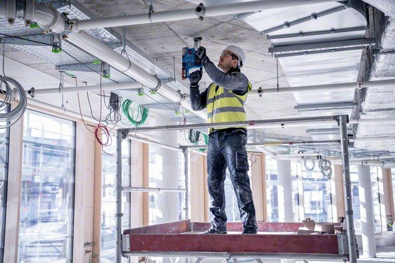 Dělník v reflexní vestě a ochranné přilbě stojí na lešení a provádí instalaci stropu v budově.