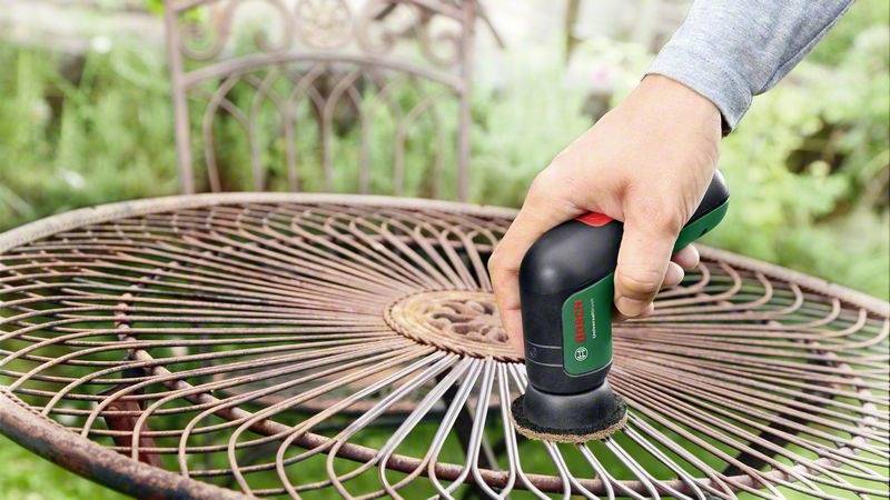 Osoba používá praktický elektrický brusný přístroj k odstranění rzi z kovového stolu venku.