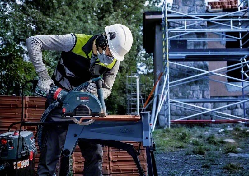 Dělník v ochranné přilbě a roušce řeže elektickou pilou dřevo na staveništi, v pozadí je lešení.
