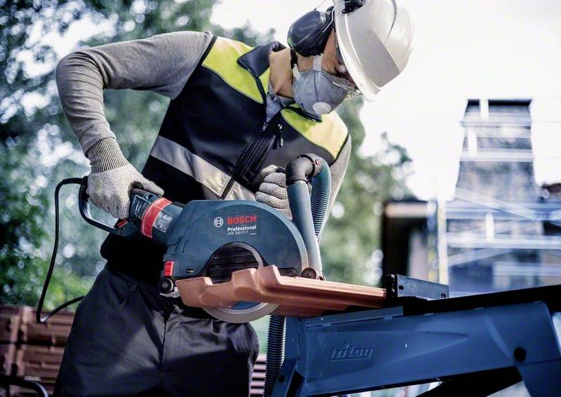 Dělník v ochranném oblečení řeže kus kovu ruční pilou Bosch. V pozadí jsou vidět stromy a lešení.