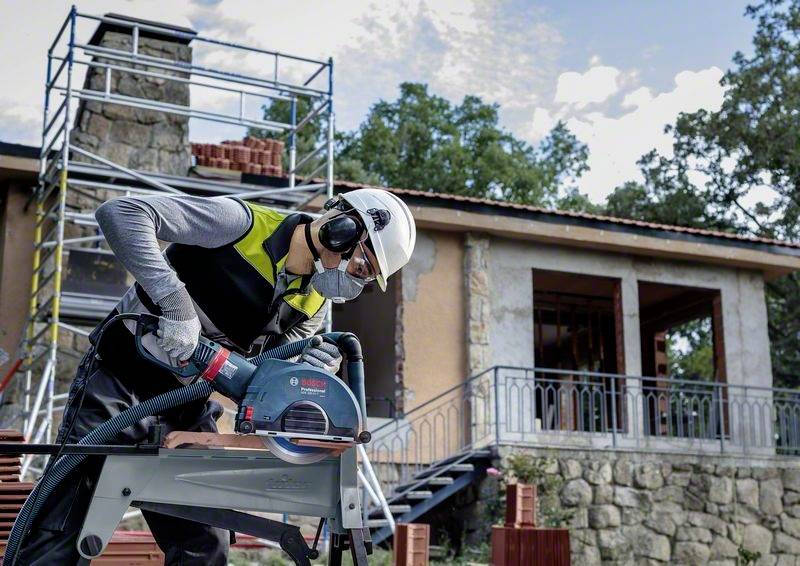 Dělník v ochranné přilbě a chráničích sluchu řeže dřevo elektrickou pilou na staveništi před budovou.