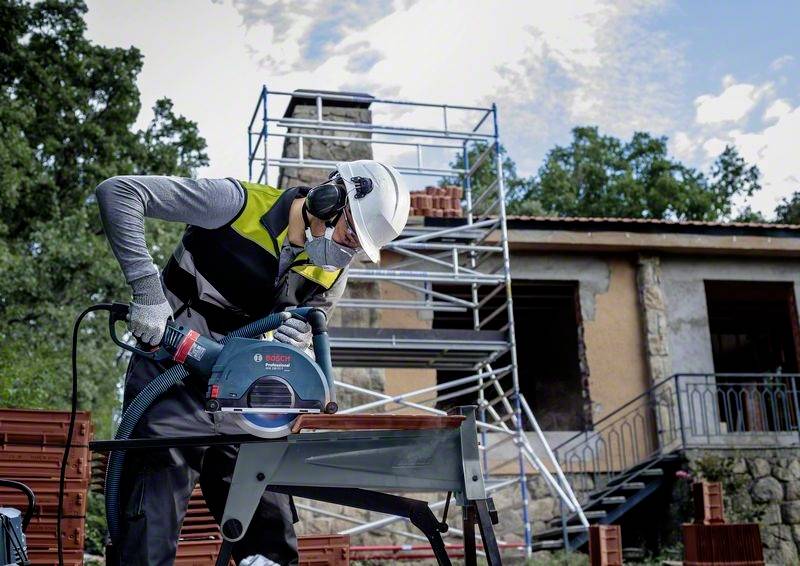Jedna osoba pracuje na staveništi s elektrickou pilou. Za ní se nachází lešení a cihlové zdivo.