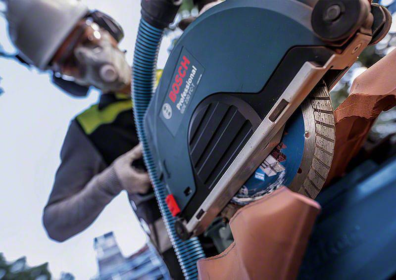 Osoba v ochranných brýlích a respirátoru řeže cihlovou střechu elektrickým pilou. Na pile je viditelný firemní název 'Bosch'.