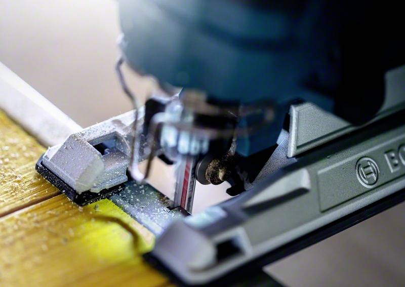 Detailní záběr elektrické přímočaré pily, která přesně řeže dřevěnou desku. Jsou viditelné piliny, ostří a detaily nástroje.