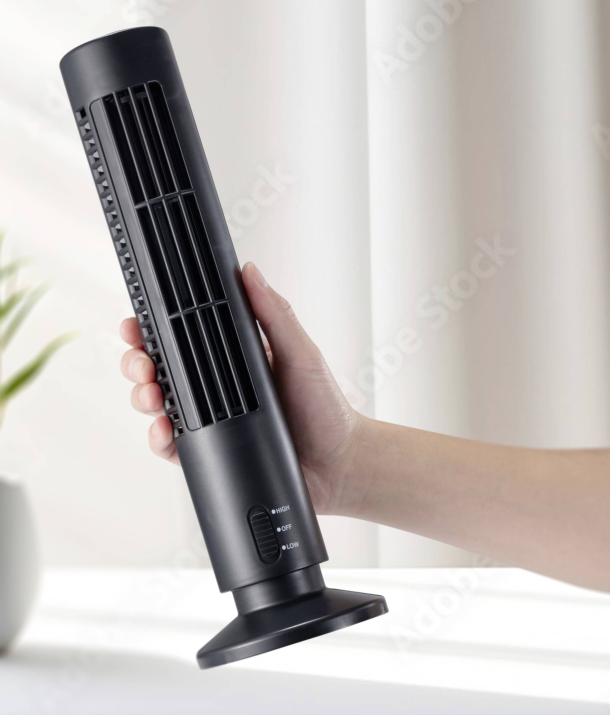 Ruka drží štíhlý černý stolní ventilátor před světlým pozadím. Vlevo v obraze stojí rostlina.