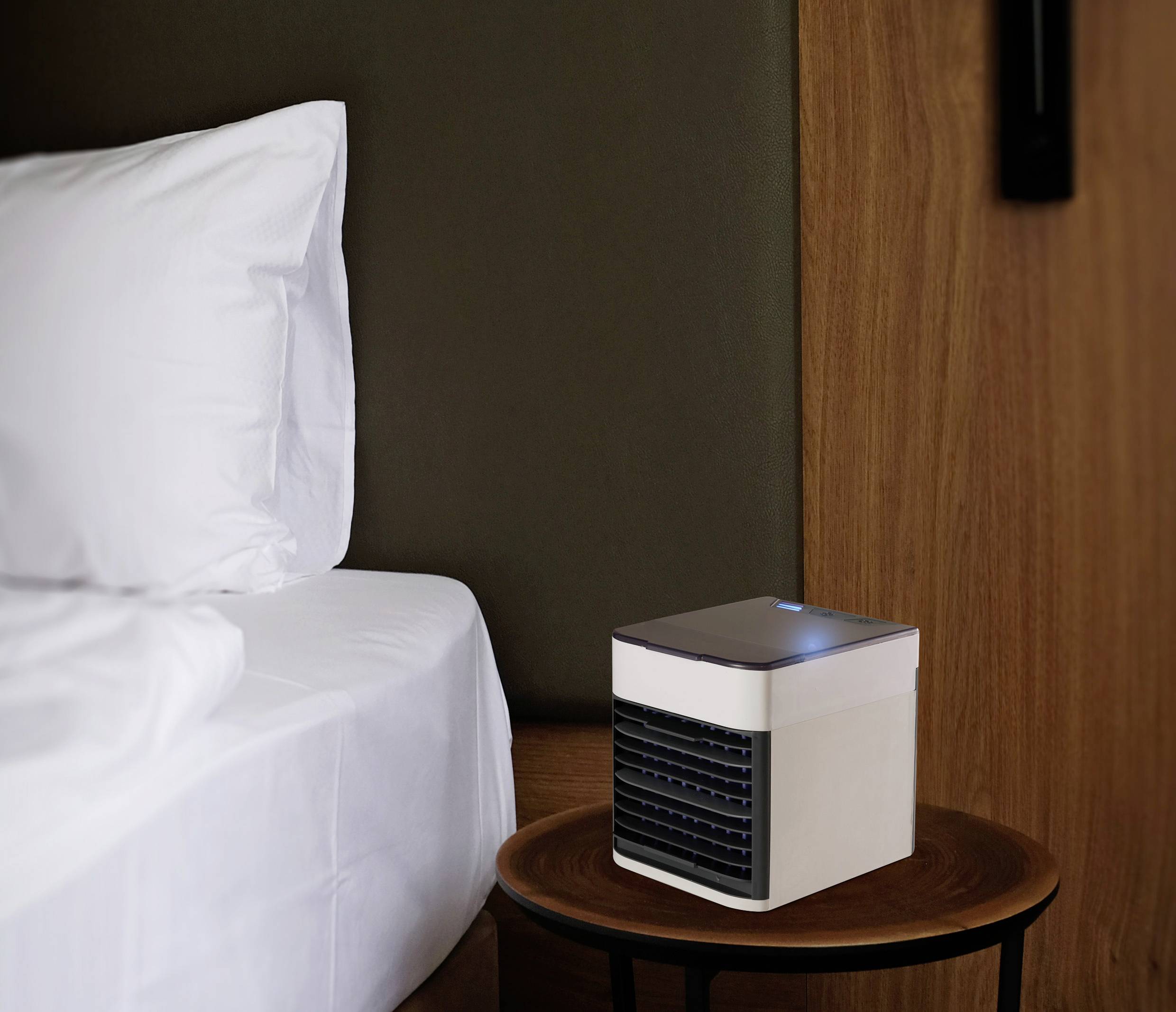 Malý bílý přenosný ventilátor je zapnutý na dřevěném stole vedle postele s bílými polštáři v moderně zařízeném pokoji.