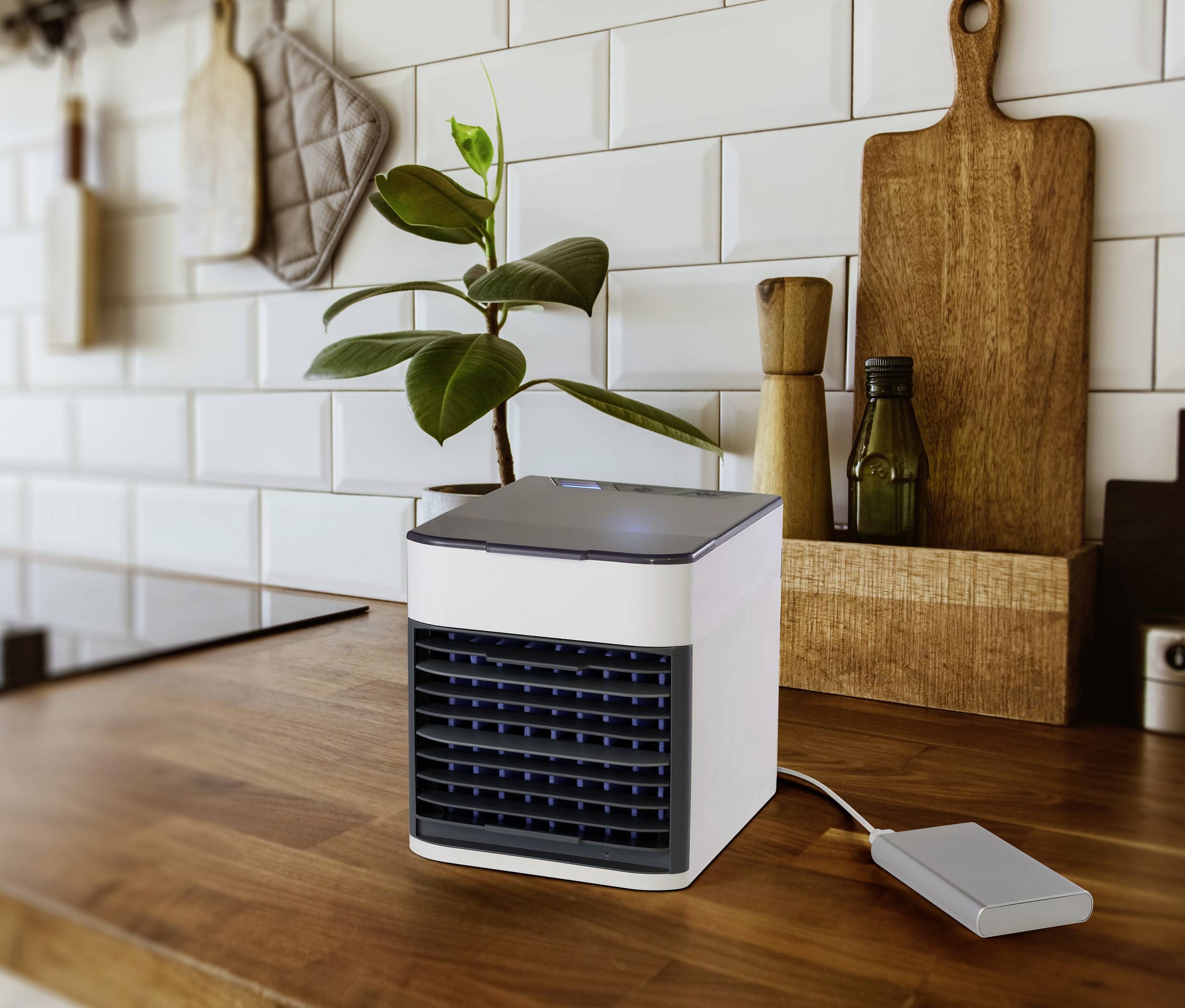 Malý přenosný klimatizační ventilátor stojí na dřevěné pracovní desce v kuchyni. Vedle něj jsou kuchyňské pomůcky a pokojová rostlina.
