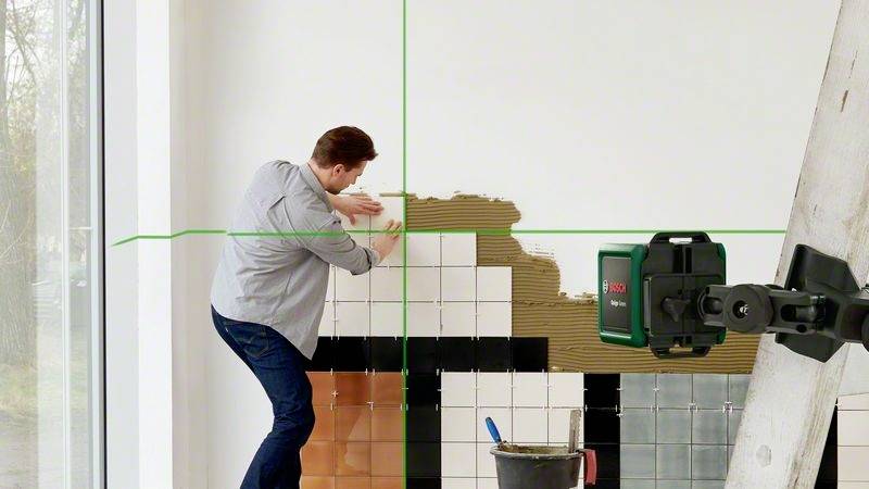 Muž upevňuje dlaždice na stěnu. Laserová pomůcka promítá zelené čáry pro správné umístění. Vedle něj stojí žebřík.