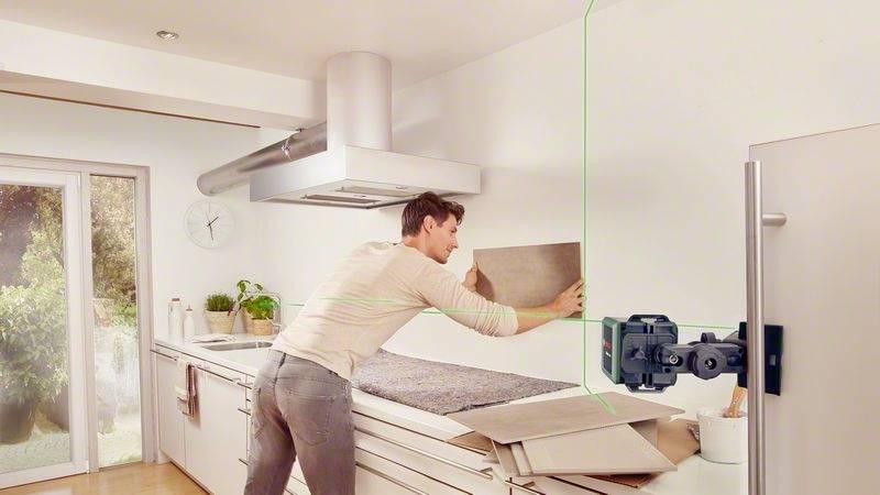Muž zaměřuje pomocí laserového zaměřovače obklad na stěně v moderní kuchyni. V pozadí je vidět velká lednice.