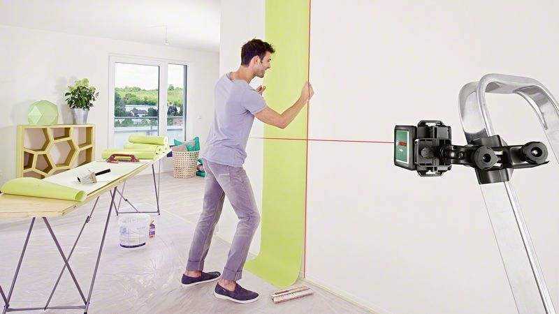Muž tapetuje stěnu světlým tapetovým pruhem v moderní místnosti. Laserový přístroj pomáhá s přesným vyrovnáním.