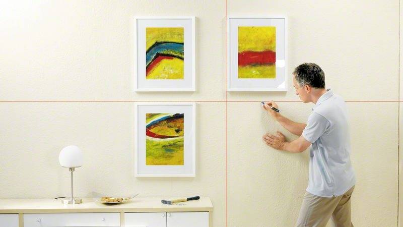 Muž používá laserovou vodováhu, aby pověsil obrázky na stěně rovně. V pozadí jsou vidět tři barevné rámy na fotografie.