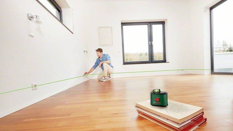 Muž používá laserový nástroj k vyznačení čáry na stěně v prázdné místnosti s dřevěnou podlahou.