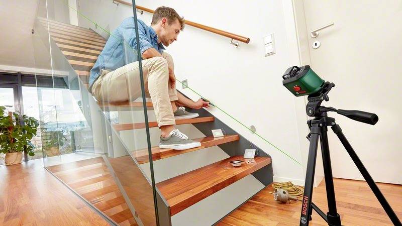 Muž používá měřicí přístroj k měření schodů dřevěného schodiště. Vedle stojí stativ.