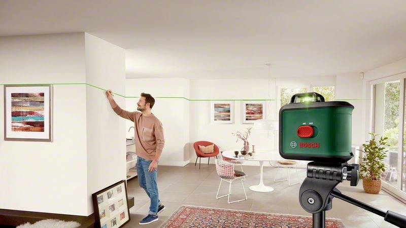 Muž zaměřuje přímou linii laserovým nivelačním přístrojem v moderním obývacím pokoji. V pozadí jsou umělecká díla.