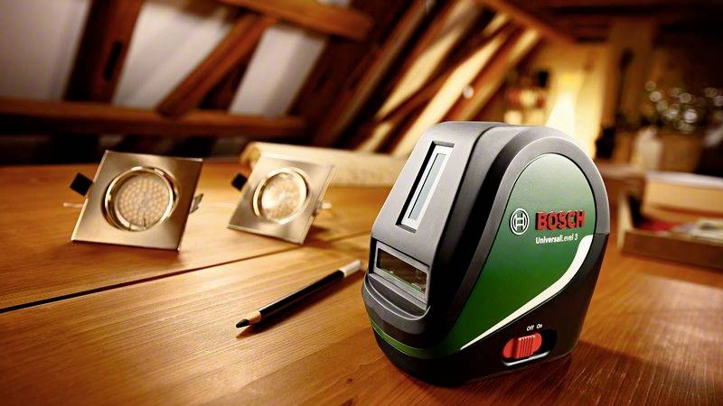 Laserový dálkoměr na dřevěném stole, vedle něj dvě vestavěné svítilny a tužka. V pozadí je nakloněný dřevěný trám.