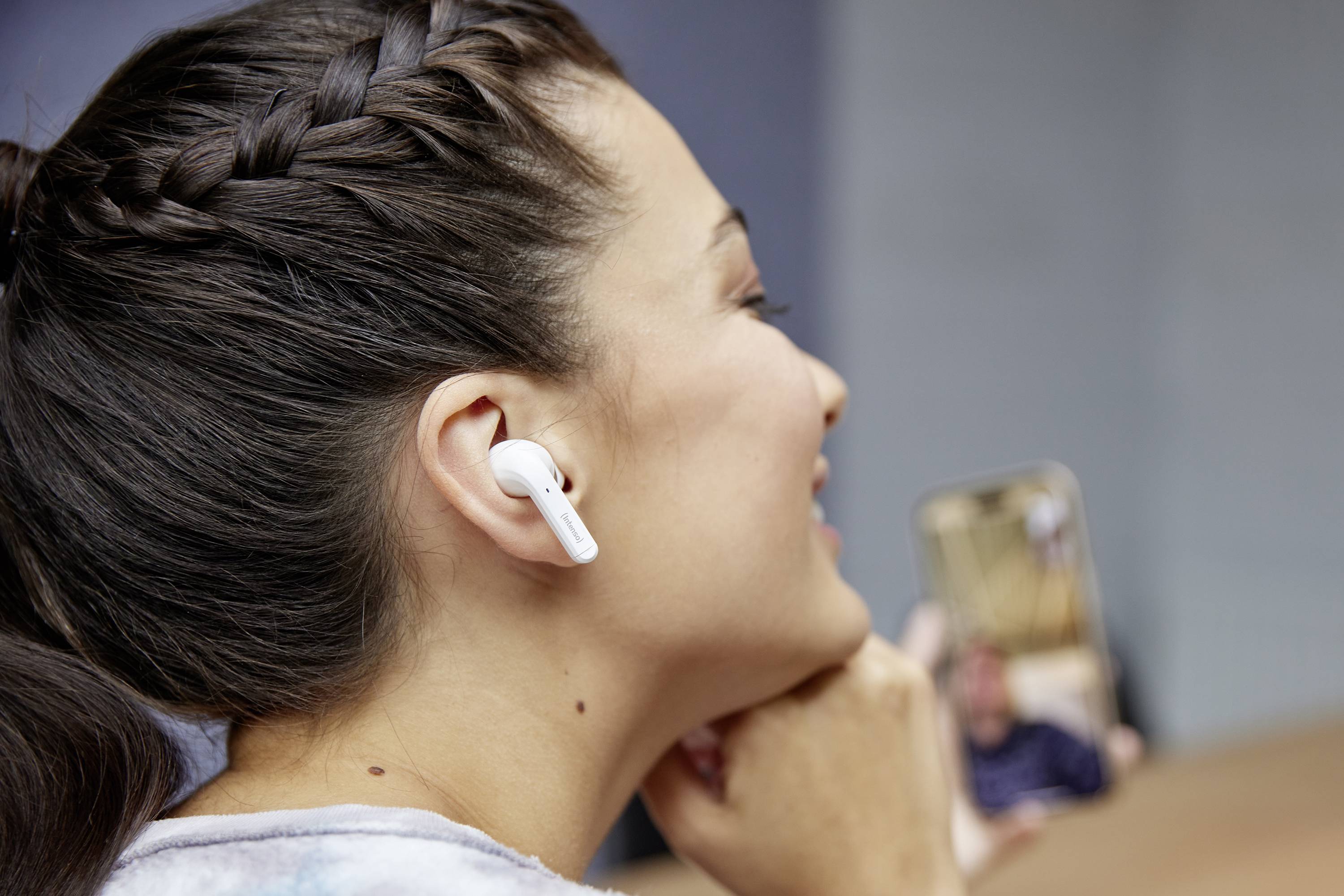 Žena s bílými bezdrátovými sluchátky provádí videohovor. Usmívá se a drží v ruce smartphone.