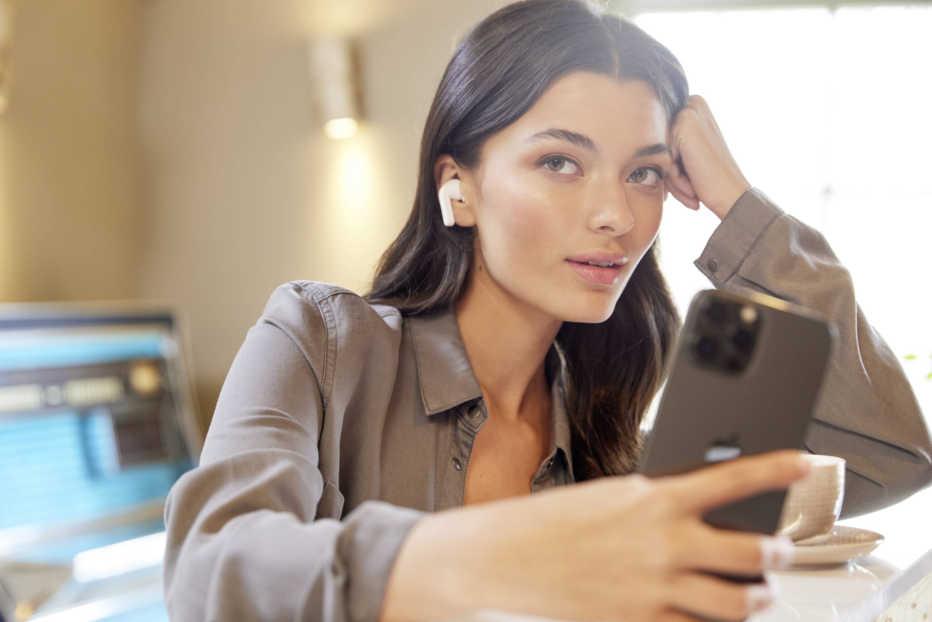 Osoba sedí u stolu, drží smartphone a má nasazená bezdrátová sluchátka. V pozadí je vidět počítačový monitor.