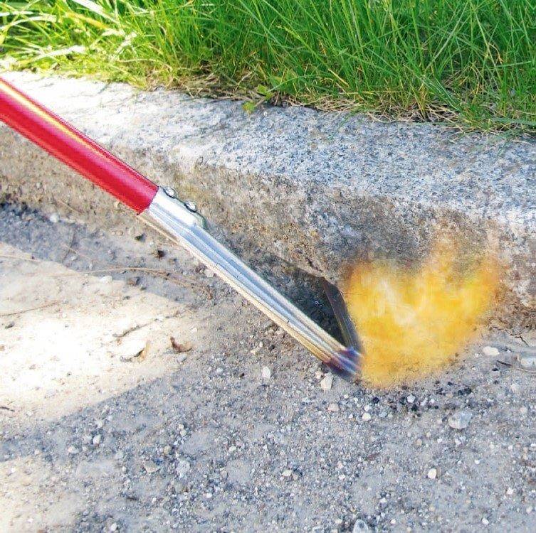 Plynový hořák s červeným držadlem se používá k odstranění plevele na okraji chodníku. Je viditelný proud plamene.