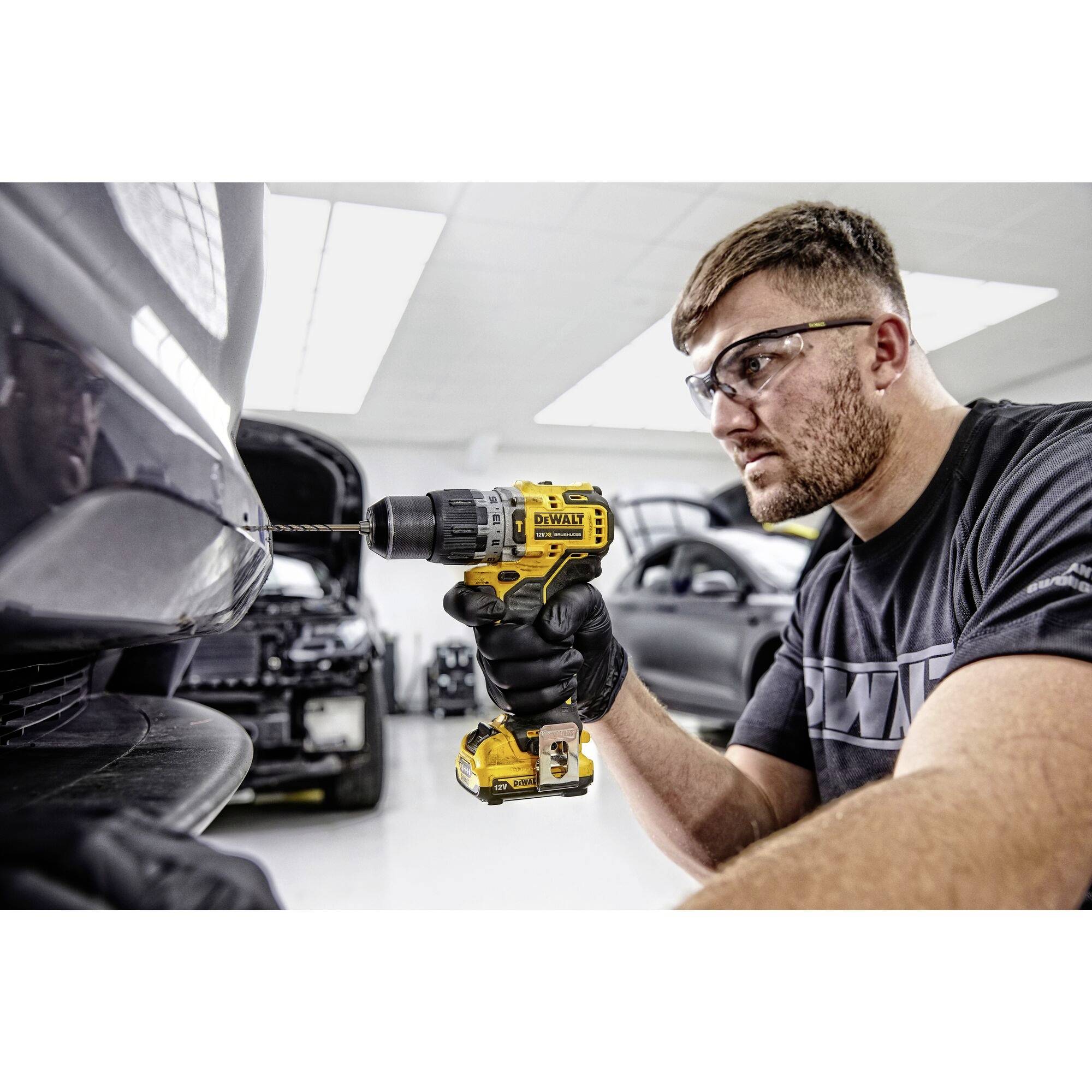Muž v ochranných brýlích vrtá žlutou vrtačkou do karoserie automobilu v prostředí dílny.