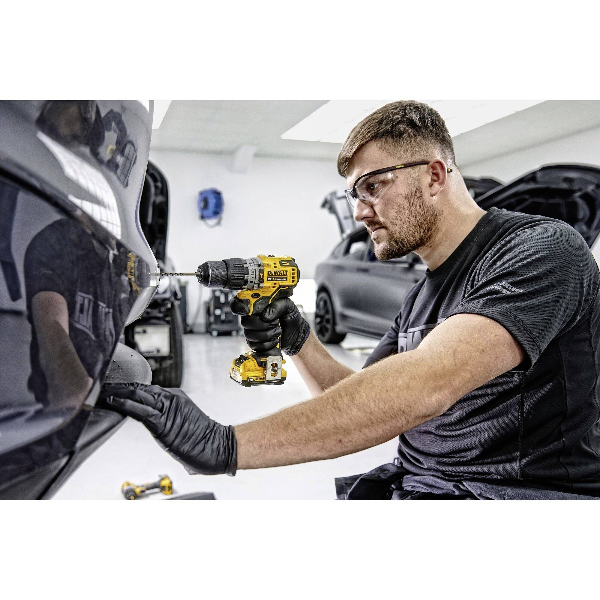 Mechanik v ochranných brýlích používá akumulátorový šroubovák při práci na automobilu. V dílně je vidět několik dalších aut.
