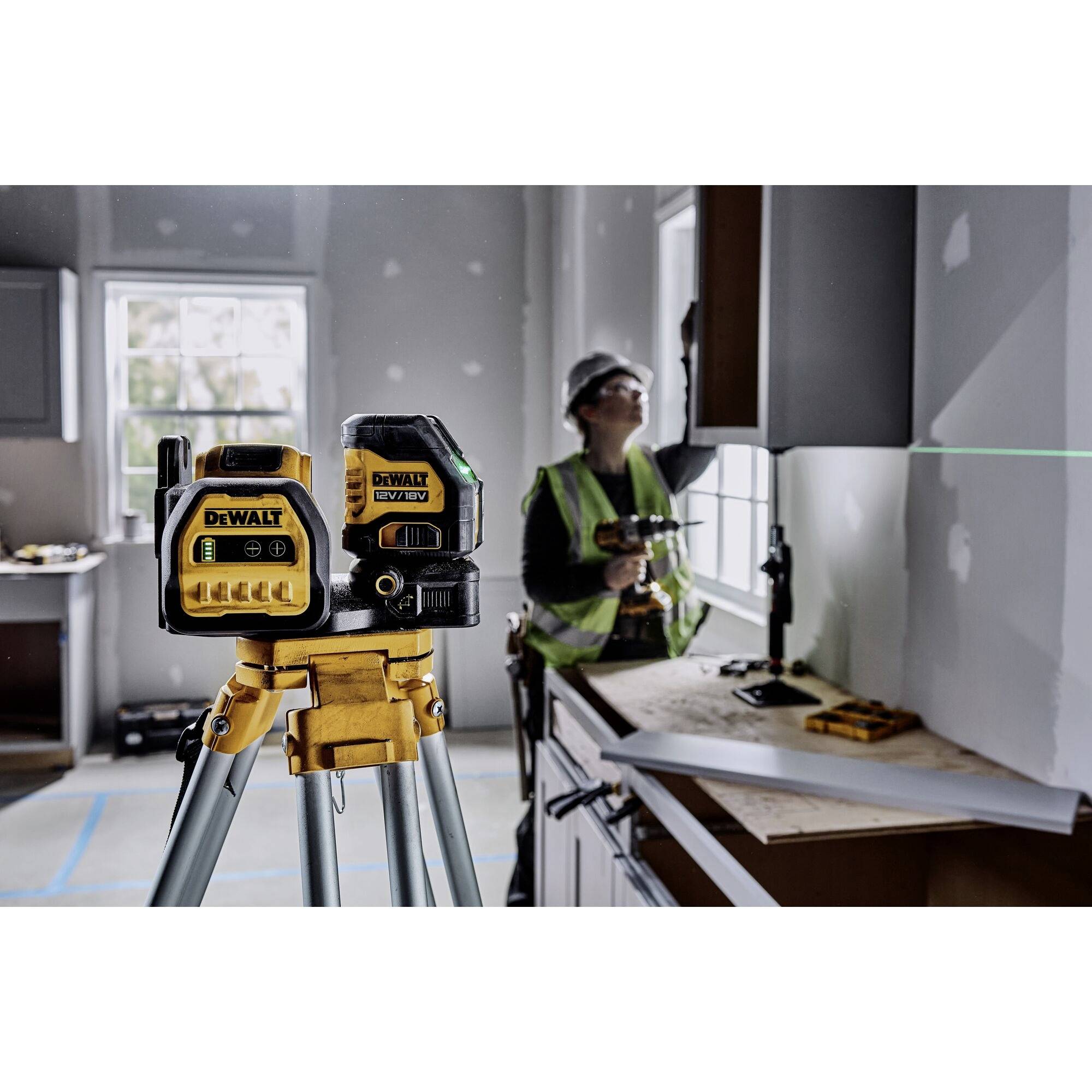 Dva laserové nivelační přístroje Dewalt stojí na stativu v místnosti. V pozadí pracuje osoba v ochranné přilbě u zdi.