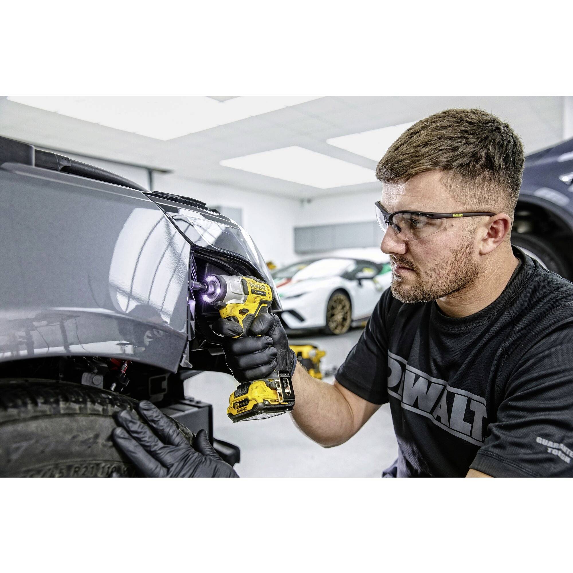Muž v ochranných brýlích opravuje nárazník automobilu v dílně pomocí akumulátorového šroubováku.