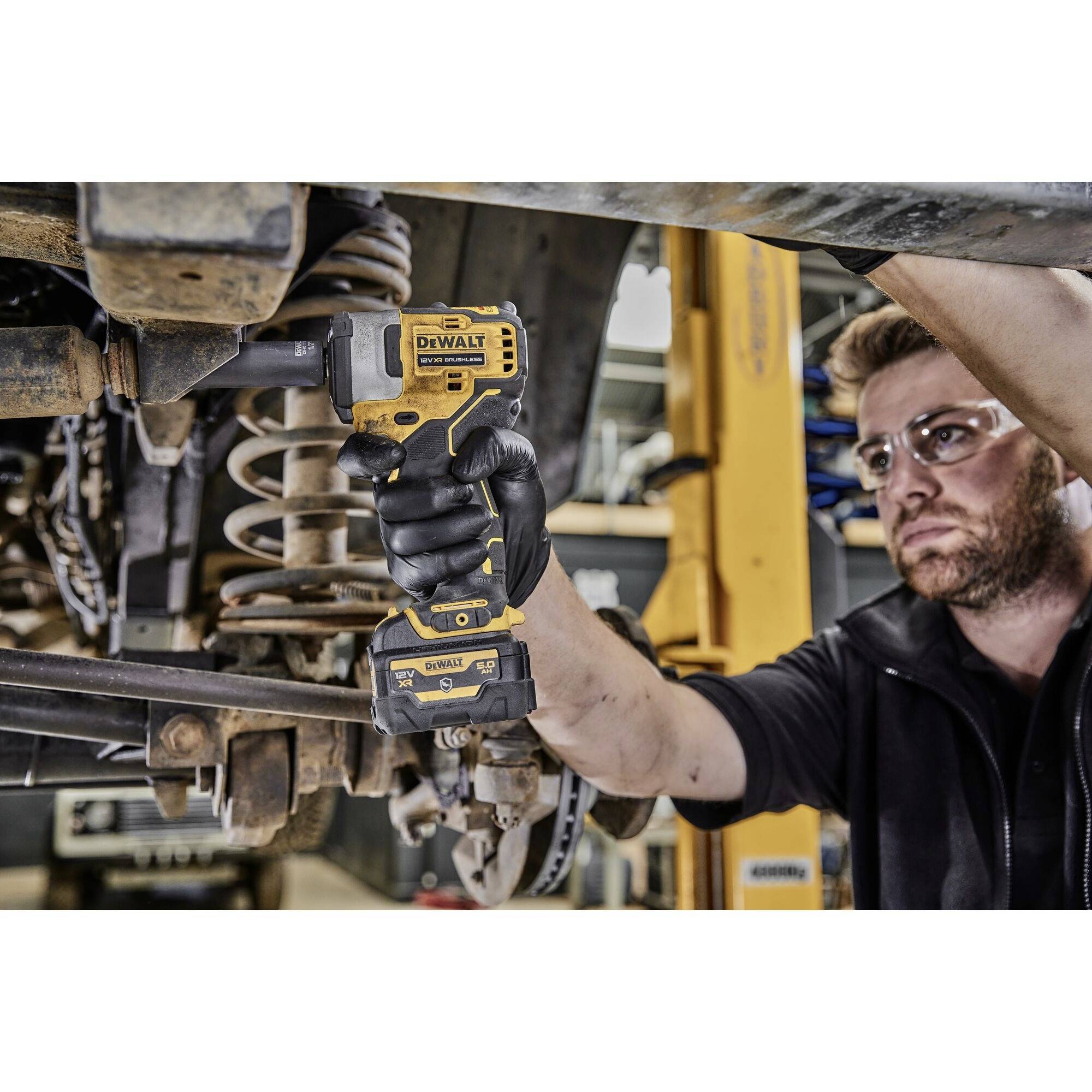 Mechanik používá žlutý akumulátorový šroubovák při práci na vozidle. V pozadí je vidět zvedák.