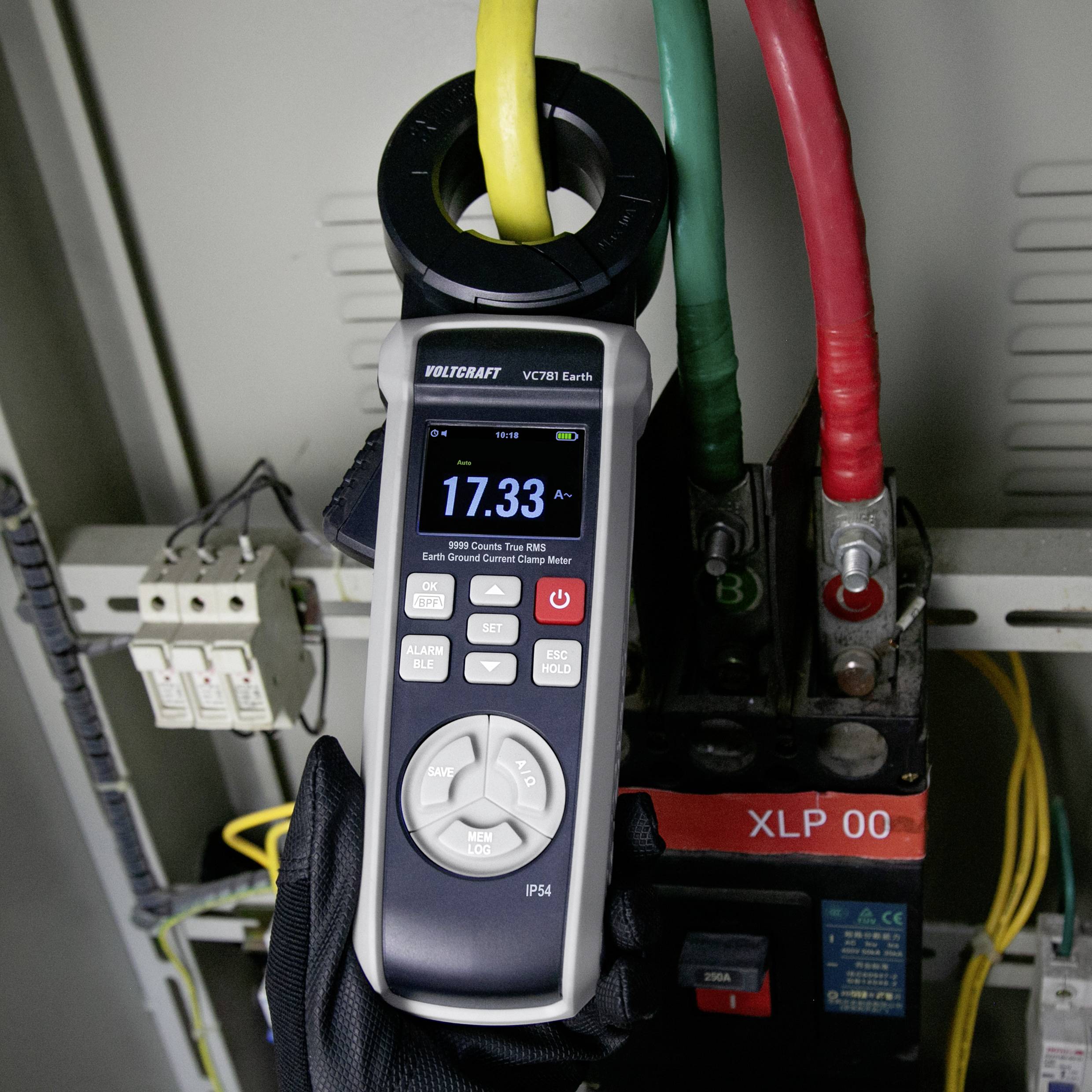 Ruka držící digitální klešťový měřič, měřící únikový proud v elektrickém rozvaděči, s viditelnými vodiči a jističem.
