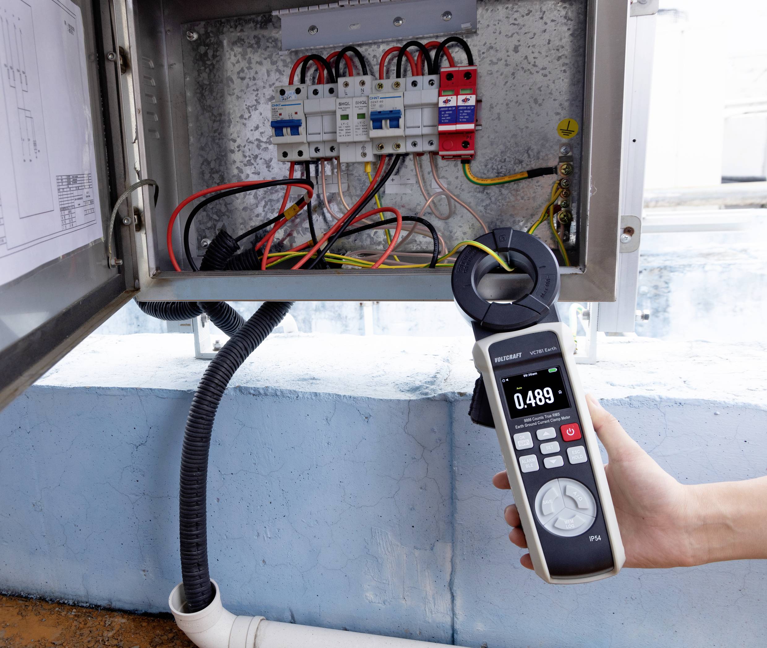 Ruka držící digitální multimetr zobrazující hodnotu 0,489 poblíž elektrického rozvaděče s viditelnými vodiči a jističi, modrá stěna v pozadí.