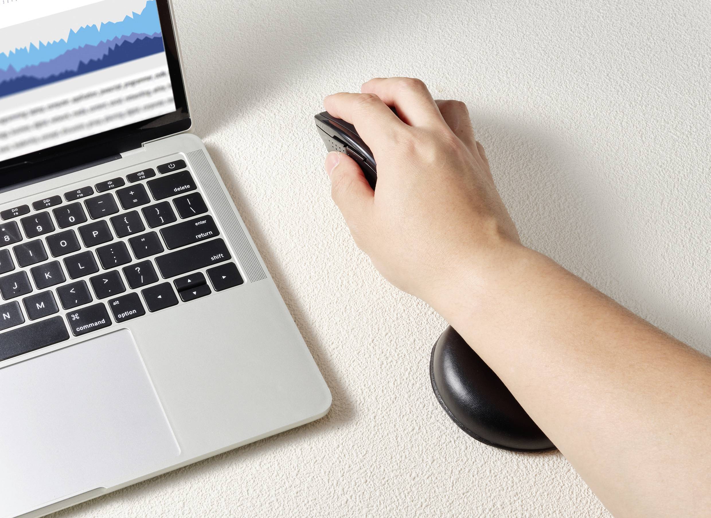 Ruka držící počítačovou myš na ergonomickém podložce pod myš; vedle notebook s diagramem na obrazovce.