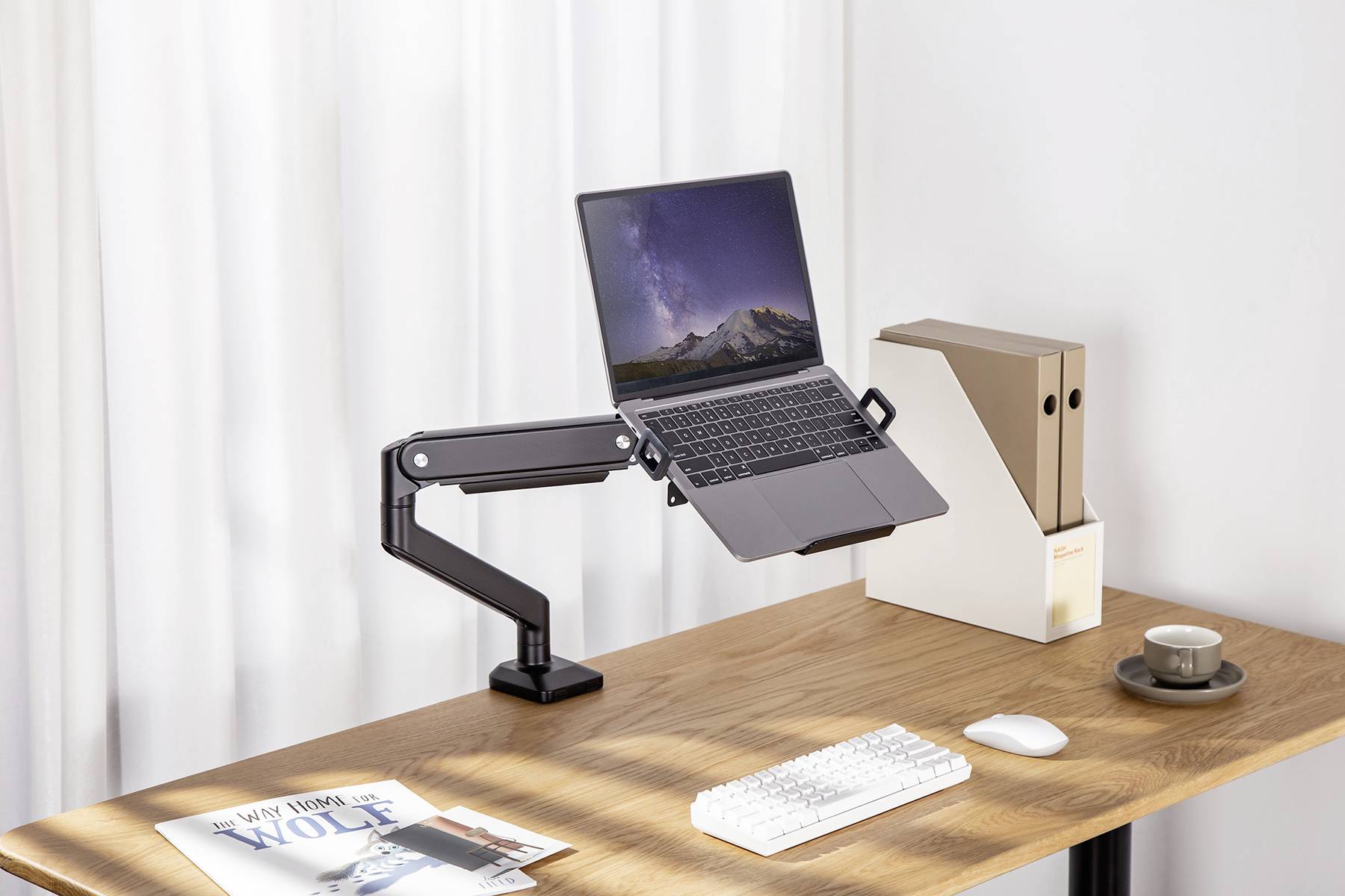 Laptop stojí na nastavitelném rameni na psacím stole. Vedle něj leží dvě složky, šálek a časopis.