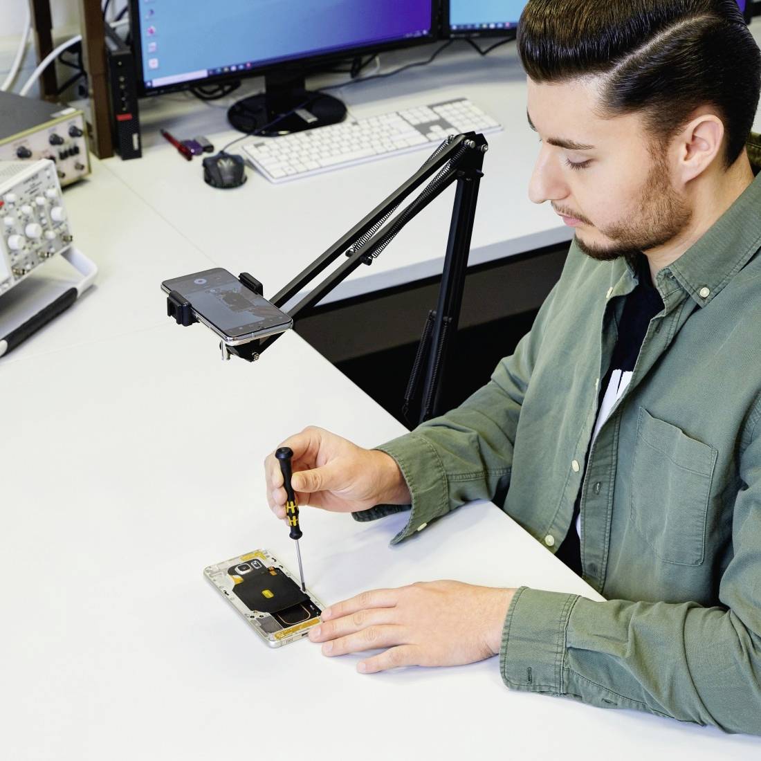 Muž opravuje smartphone na stole s technickým vybavením, včetně mikroskopu a specializovaných nástrojů.