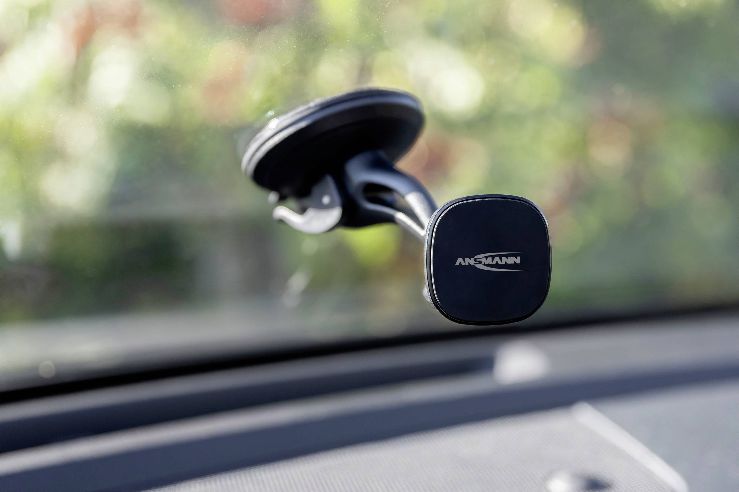 Magnetická držák na mobil od značky Ansmann upevněný na čelním skle automobilu, v pozadí rozmazaná krajina.