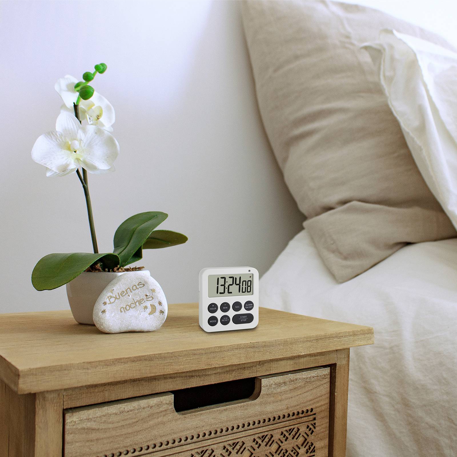 Digitální budík ukazuje 13:42 na nočním stolku vedle bílé orchideje v květináči s nápisem 'buenos días'.