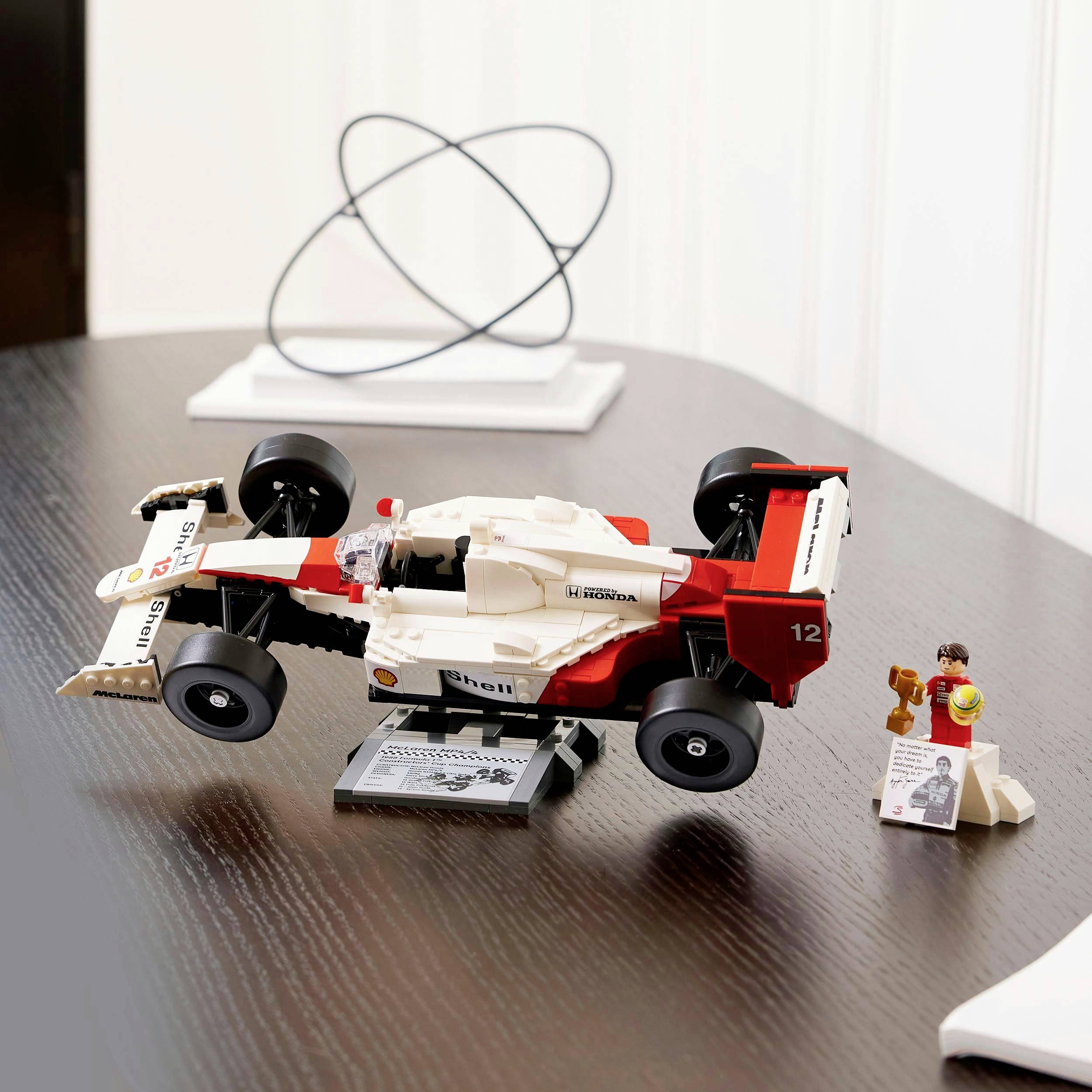 Model auta LEGO s nápisem 'Honda' a číslem 12 stojí na stole. Vedle něj je figurka s helmou.