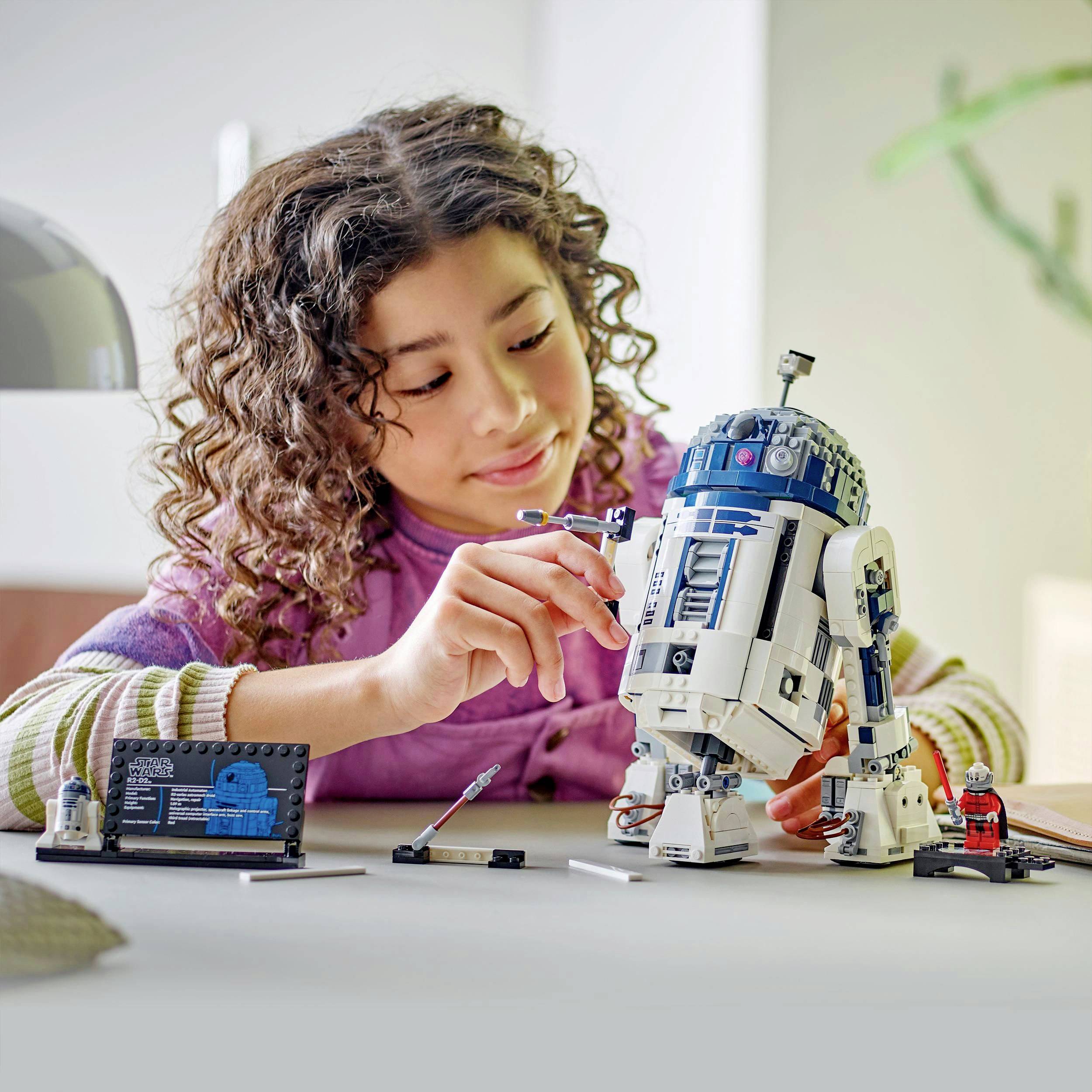 Dívka staví model R2-D2 u stolu, obklopena díly Lega, s velmi soustředěným výrazem ve tváři.