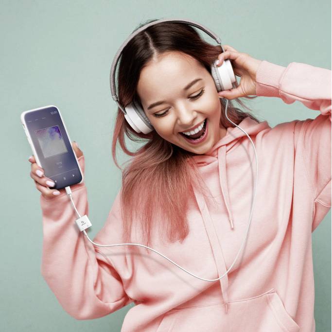Usměvavá osoba s nasazenými sluchátky poslouchá hudbu a drží smartphone. Má na sobě růžovou mikinu s kapucí.