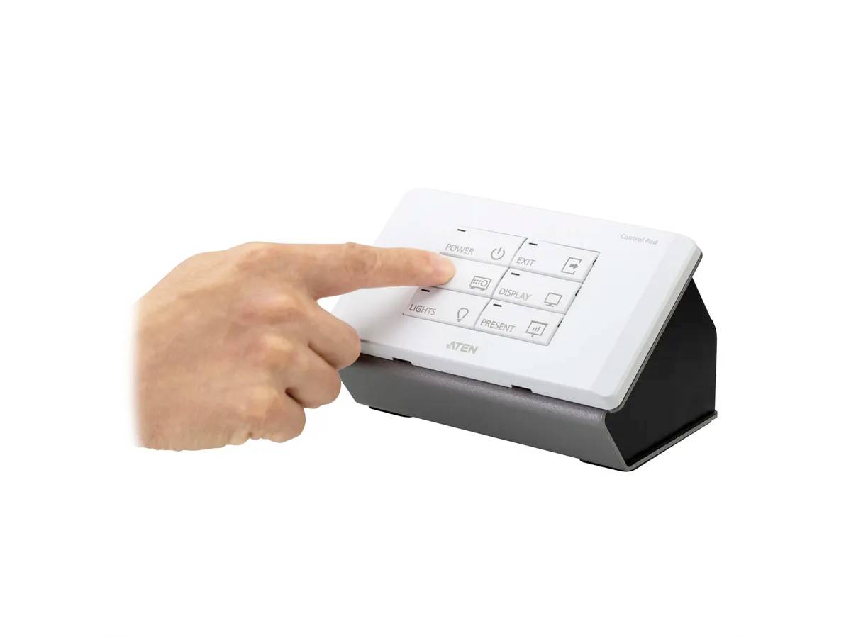 Jedna ruka ukazuje na elektronický ovládací panel s několika tlačítky. Panel je umístěn na stole nebo jiné ploše.