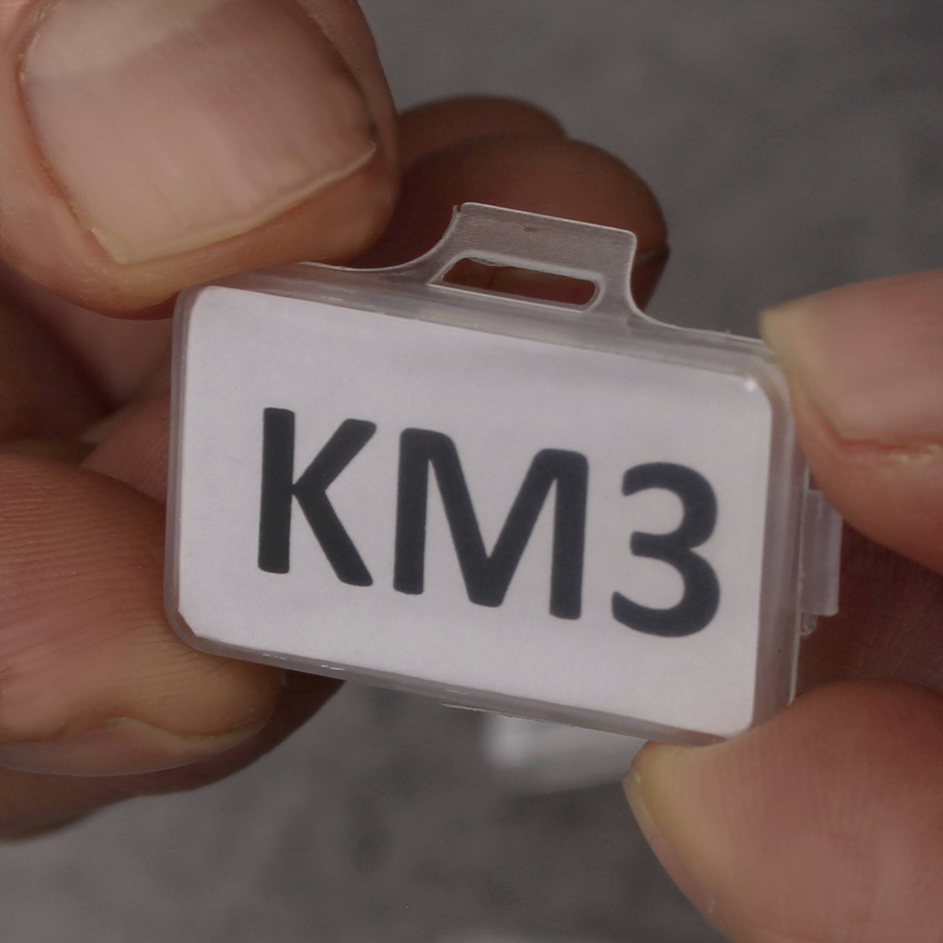 Ruka drží malou plastovou nálepku s nápisem 'KM3'.