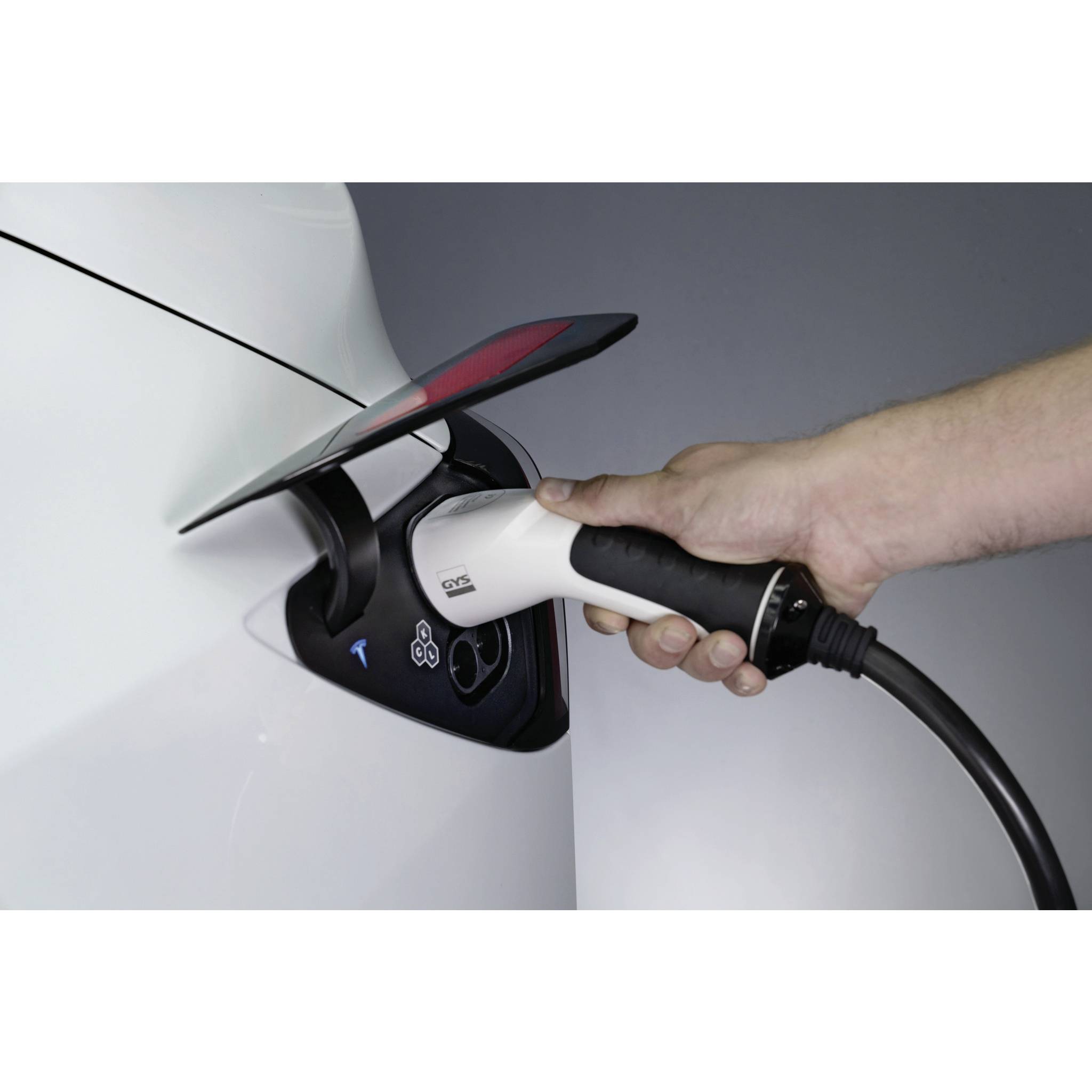 Ruka drží nabíjecí kabel, který je připojen k bílému elektromobilu. Vozidlo je právě nabíjeno.