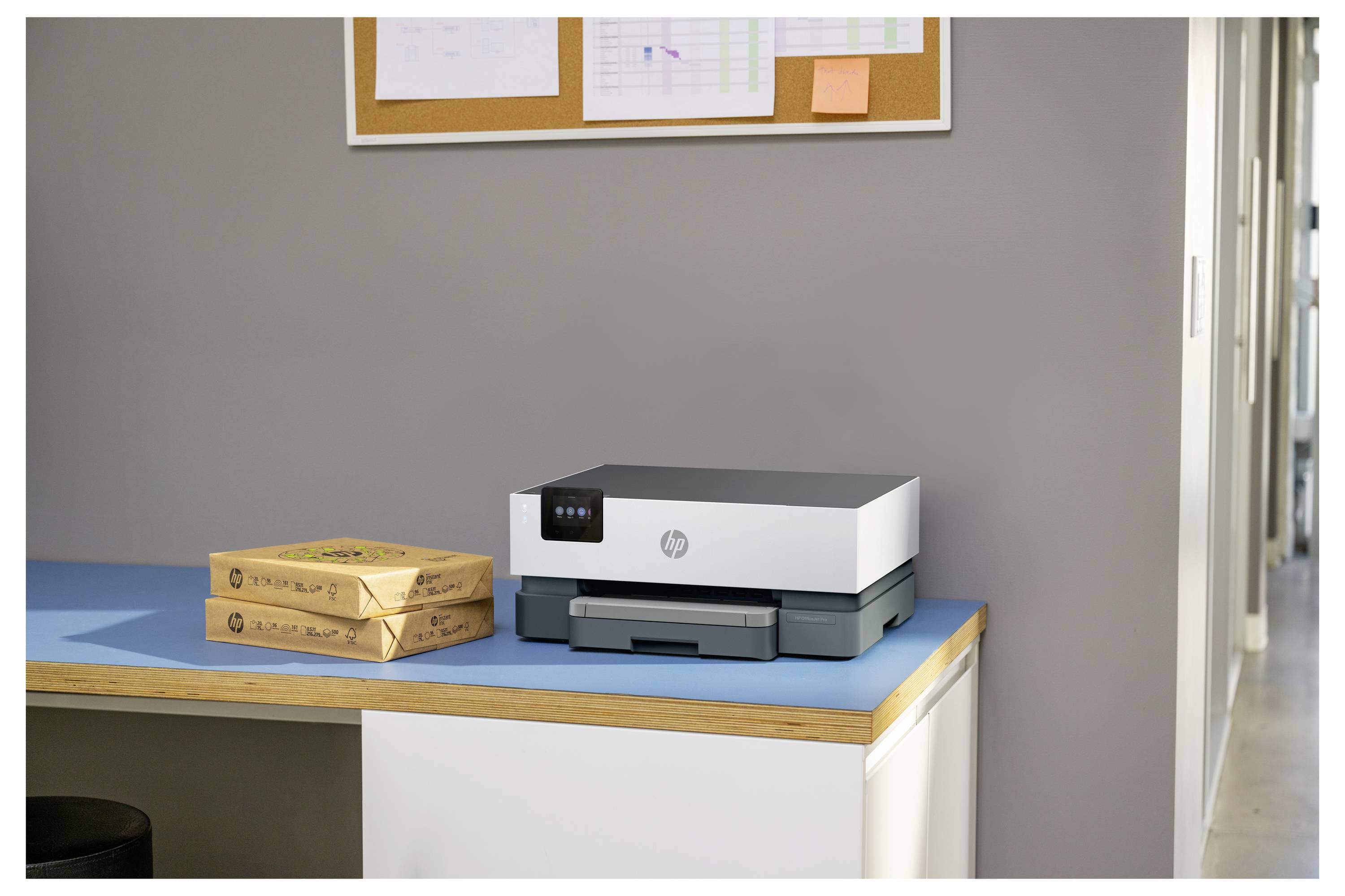 Malá kancelářská stůl s tiskárnou HP vedle dvou naskládaných kartonových poštovních krabic, což naznačuje zasílání balíků nebo tisk etiket.