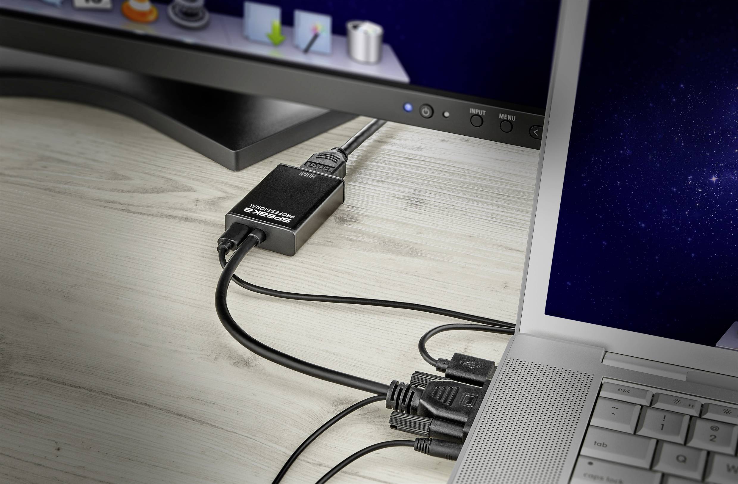 Notebook připojený k desktopovému monitoru. Několik kabelů vede do adaptéru, který umožňuje připojení. Scéna pracovního místa.