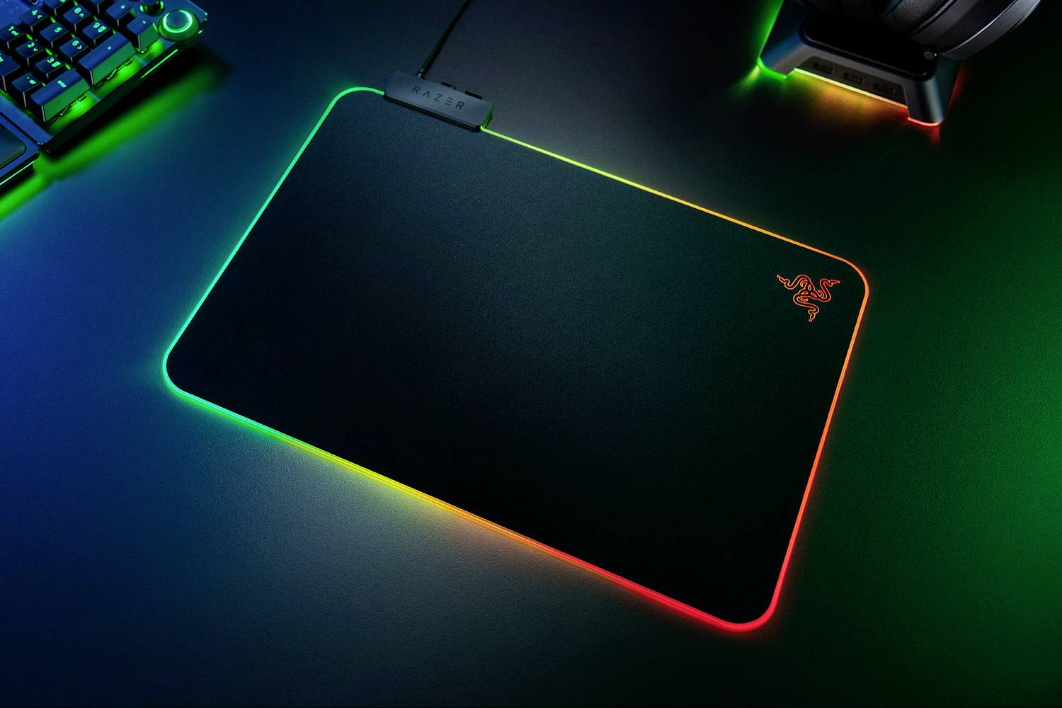 Zářící podložka pod myš s RGB podsvícením leží na psacím stole, obklopená klávesnicí a sluchátky se stejně zářivými akcenty.