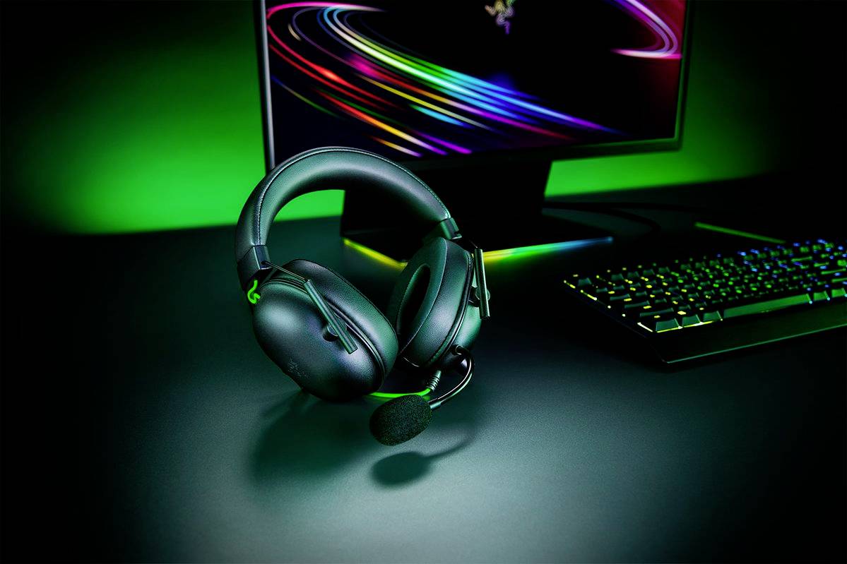 Herní headset s mikrofonem leží na stole před monitorem a podsvícenou klávesnicí. Sestava je ponořena do zelenkavého světla.