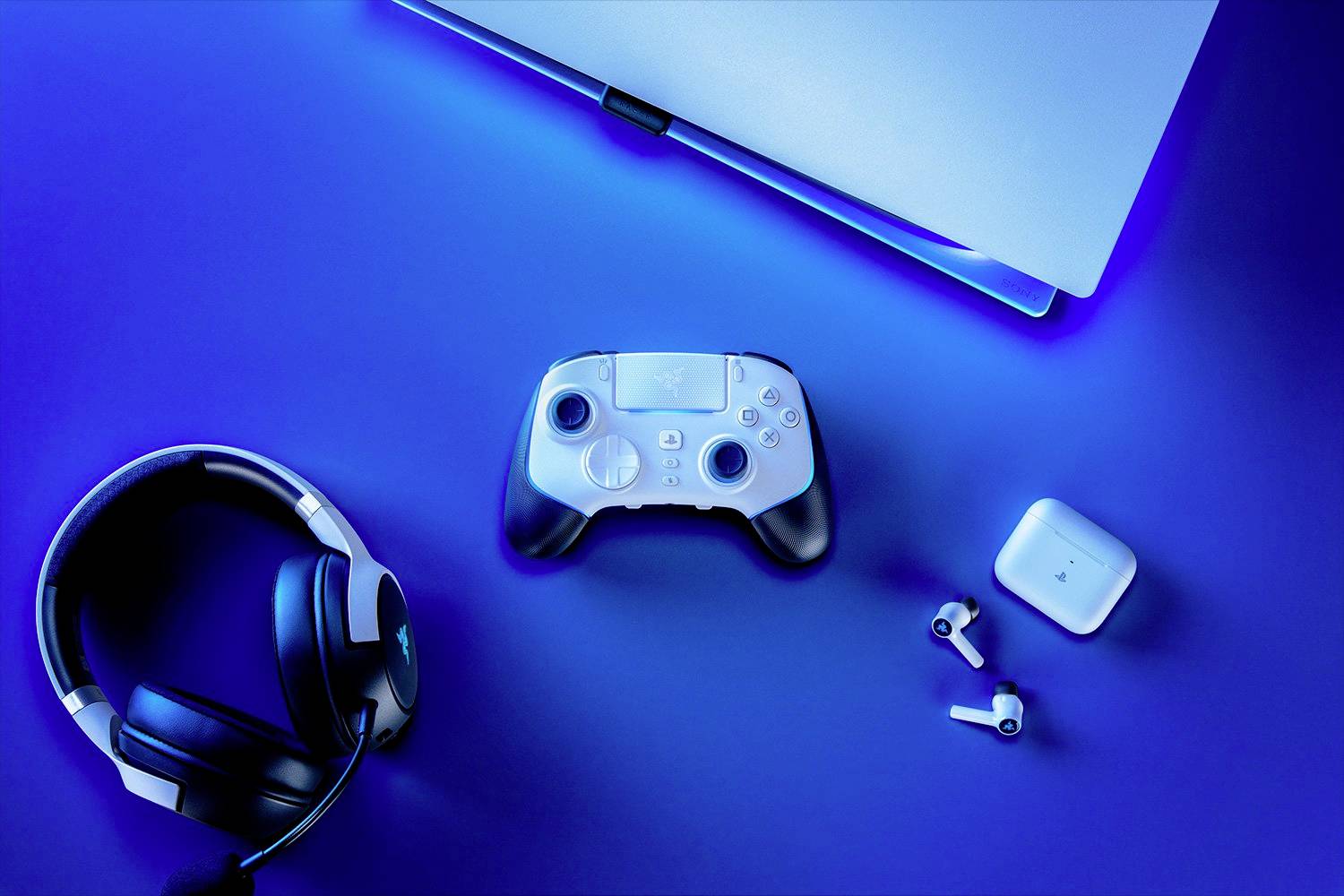 Bílý herní ovladač leží uprostřed modrého stolu. Vedle něj jsou laptop, černá sluchátka a bezdrátové earbuds.