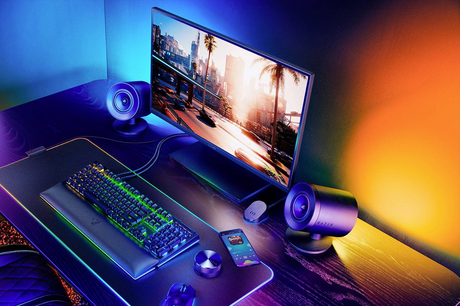 Barevně pestrý herní setup s monitorem, klávesnicí, myší a reproduktory na stole. Na obrazovce je zobrazený pohled na město.