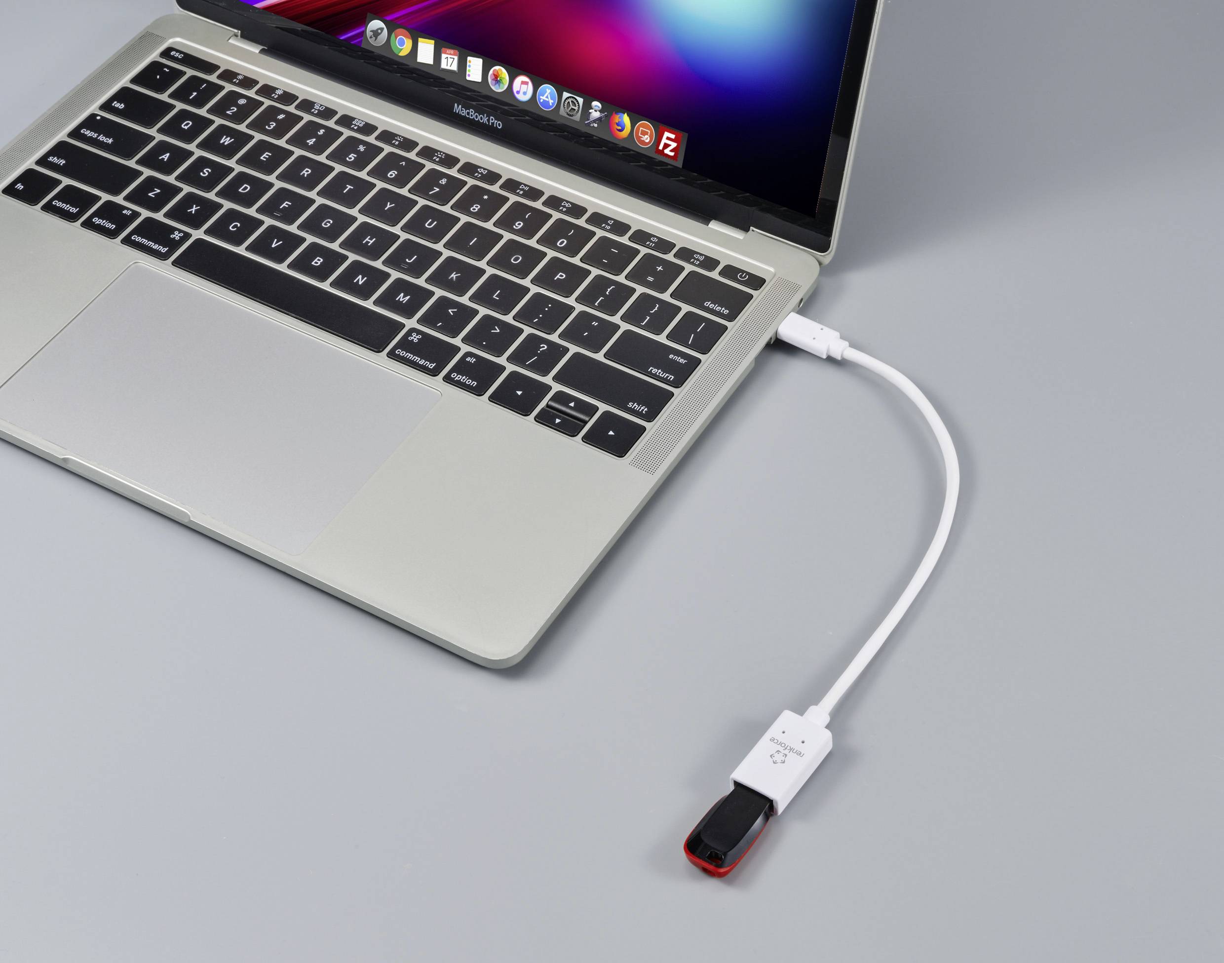 Laptop s otevřenou obrazovkou je připojen krátkým bílým kabelem k USB flash disku ležícímu na stole.