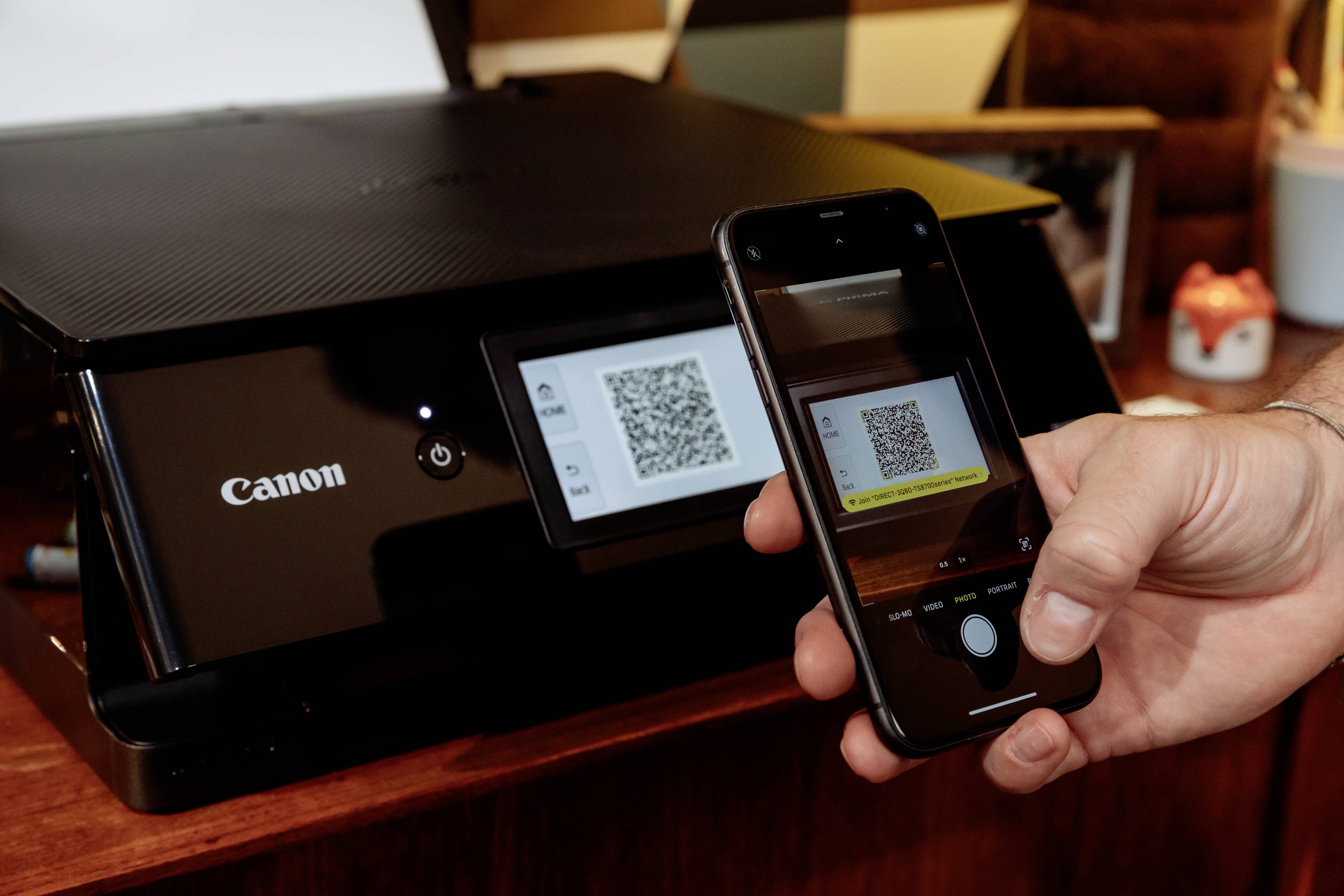 Ruka drží smartphone a skenuje QR kód na displeji tiskárny. Tiskárna v černé barvě, detailní záběr technologie v akci.