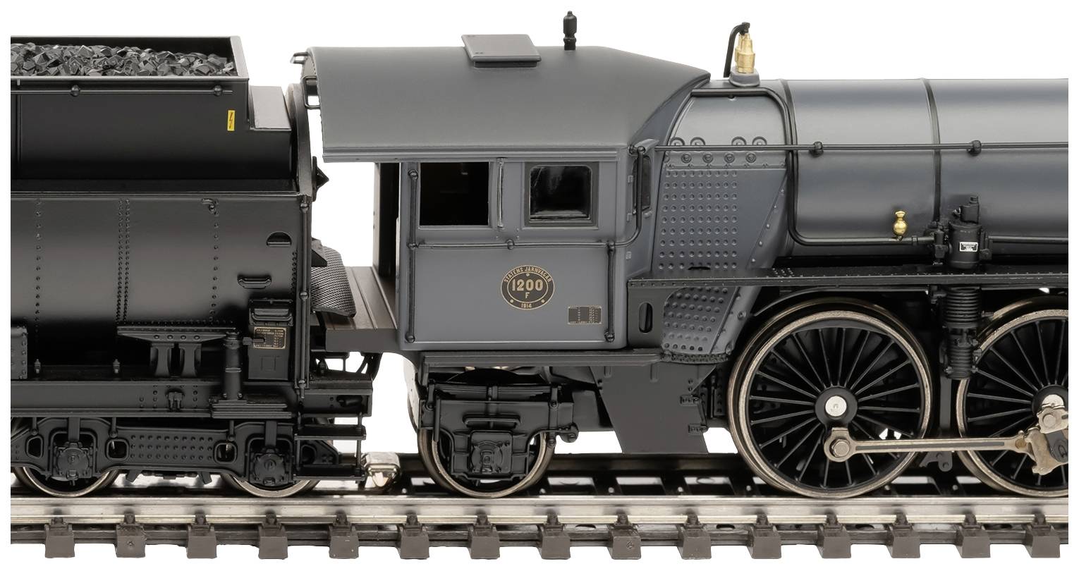Model vlaku v detailním záběru ukazuje podrobné části lokomotivy a velká kola na kolejích. Vlak je tmavě šedý a černý.